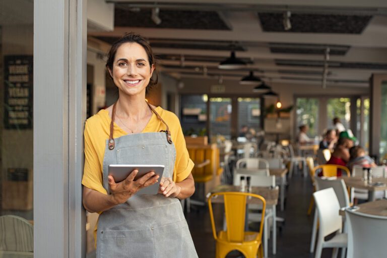 A smiling restaurant owner leaning against a pillar and working on their tablet