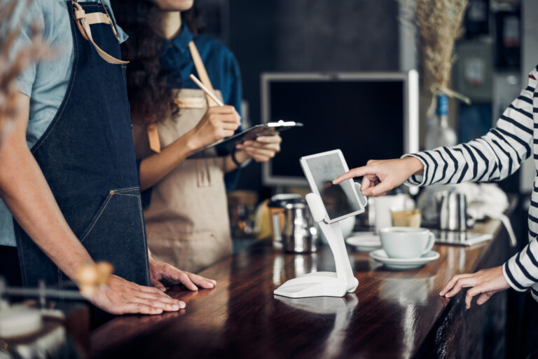 A customer selects options on a self-service tablet at a restaurant counter while two employees observe